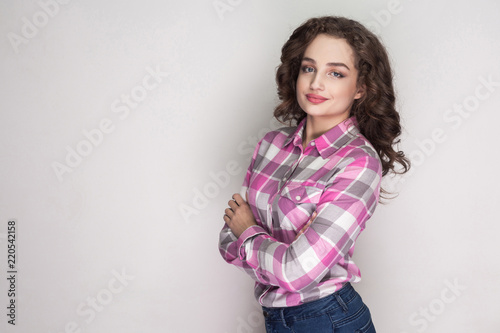 Satisfied beautiful girl with pink checkered shirt, curly hairstyle and makeup standing, crossed hands and looking at camera and smiling. indoor studio shot, isolated on gray background. © khosrork