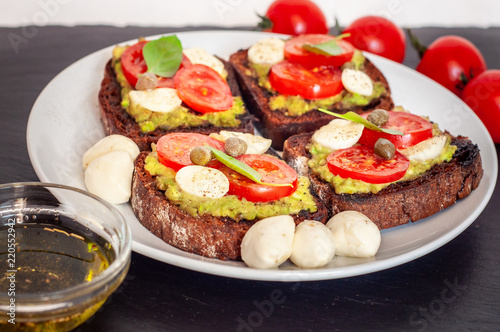 Sandwich toasts with tomatoes cherry, mozzarella, avocado, basil and olive oil. Side view on a dark stone dish.