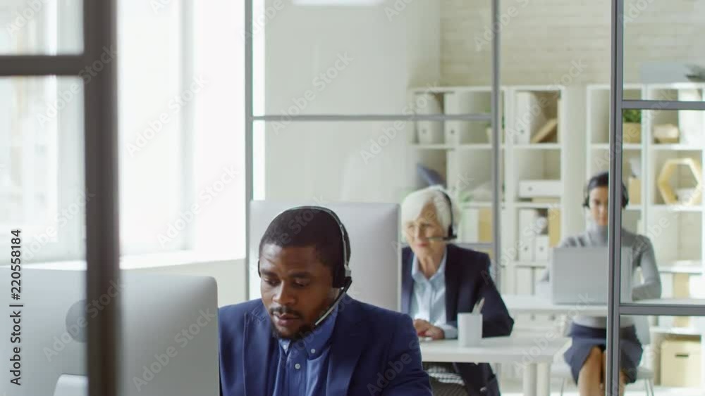 Group of multi ethnic office workers in headsets sitting at desks and using laptop and desktop computers, tilt down