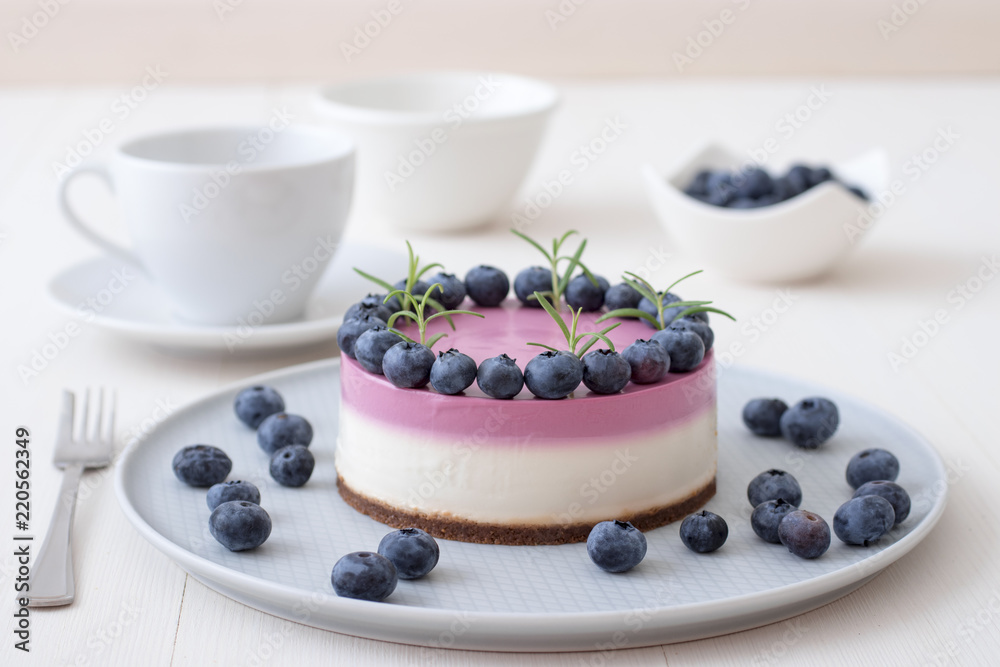 The two-color blueberry mini cheese cake. Round no bake cheesecake, bowl with blueberries, cup of coffee and bowl with sugar. Top of cake decorated by rosemary herbs. White table. The two-color blueberry mini cheese cake. Round no bake cheesecake, bowl with blueberries, cup of coffee and bowl with sugar. Top of cake decorated by rosemary herbs. White table.