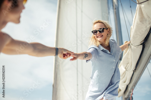 Happy woman on sailing ship stretching hand to her male parner, beautiful young carefree woman in sunglasses stands against white sails. Love smile, concept summer ocean holiday travel. photo
