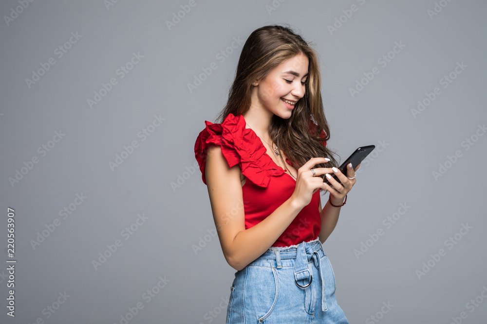Naklejka premium Attractive smiling girl standing against gray background and typing sms on her smartphone