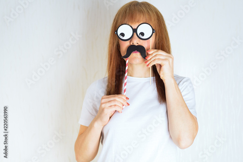 Attractive middle aged woman having fun with a fake moustache and glasses. A woman in the period of menopause.Blond / red hair mature senior female against the white wall background.Impostor syndrome