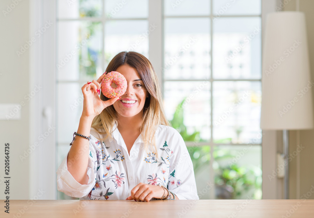 Young woman at home eating a doughnut with a happy face standing and smiling with a confident smile showing teeth Young woman at home eating a doughnut with a happy face standing and smiling with a confident smile showing teeth