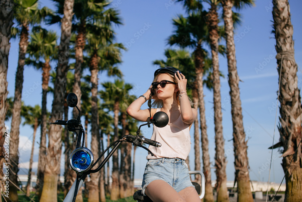 Obraz premium Cheerful female in trendy summer clothes and sunglasses sitting on electrical bicycle over city tropical palm garden background in a sunny day.