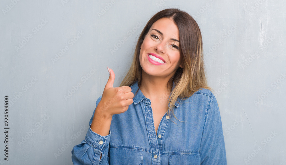 Young adult woman over grunge grey wall wearing denim outfit doing happy thumbs up gesture with hand. Approving expression looking at the camera with showing success. Young adult woman over grunge grey wall wearing denim outfit doing happy thumbs up gesture with hand. Approving expression looking at the camera with showing success.