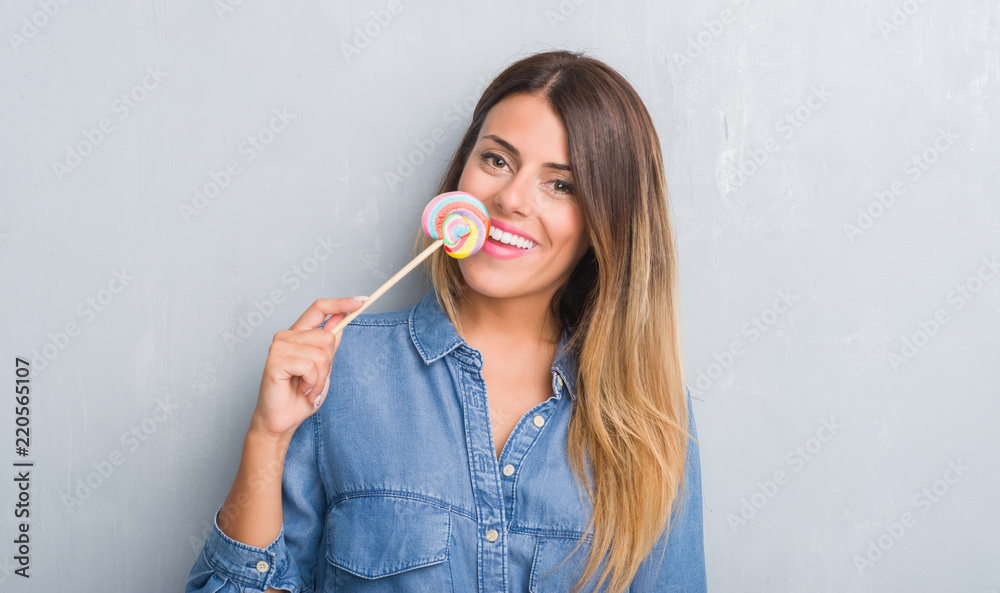 Fototapeta premium Young adult woman over grey grunge wall eating lollipop candy with a happy face standing and smiling with a confident smile showing teeth