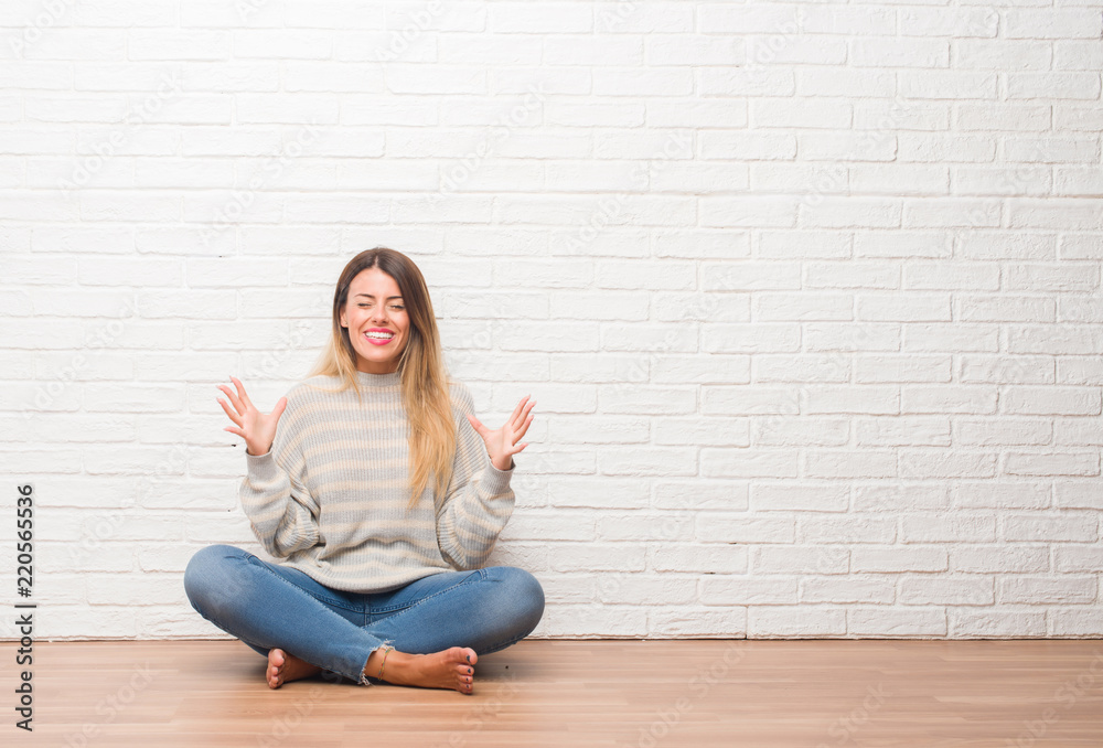 Young adult woman sitting on the floor over white brick wall at home celebrating mad and crazy for success with arms raised and closed eyes screaming excited. Winner concept Young adult woman sitting on the floor over white brick wall at home celebrating mad and crazy for success with arms raised and closed eyes screaming excited. Winner concept