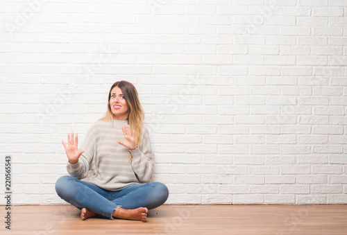 Young adult woman sitting on the floor over white brick wall at home afraid and terrified with fear expression stop gesture with hands, shouting in shock. Panic concept.