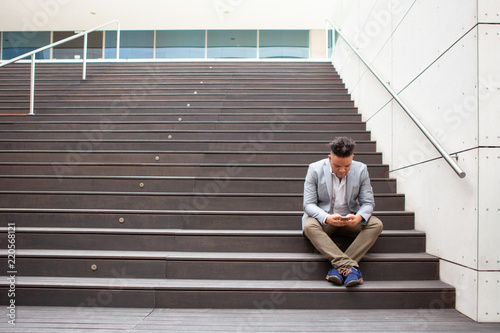 Portrait of serious businessman or student using mobile phone. Latin American young man sitting on stairs and reading or texting message. Mobile communication concept