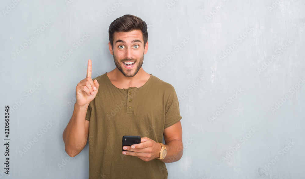 Handsome young man over grey grunge wall texting a message using smartphone surprised with an idea or question pointing finger with happy face, number one Handsome young man over grey grunge wall texting a message using smartphone surprised with an idea or question pointing finger with happy face, number one