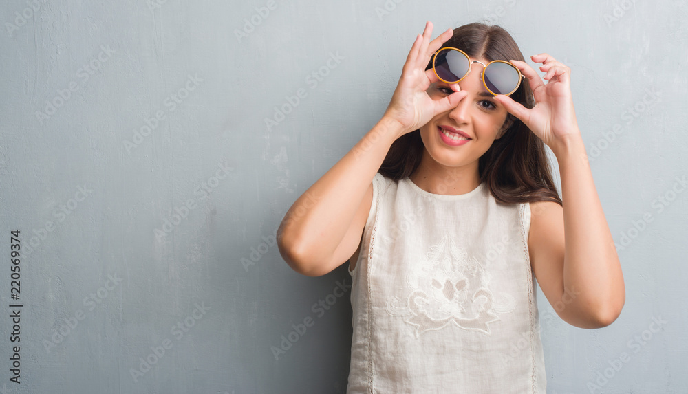 Young brunette woman over grunge grey wall wearing retro sunglasses with happy face smiling doing ok sign with hand on eye looking through fingers Young brunette woman over grunge grey wall wearing retro sunglasses with happy face smiling doing ok sign with hand on eye looking through fingers