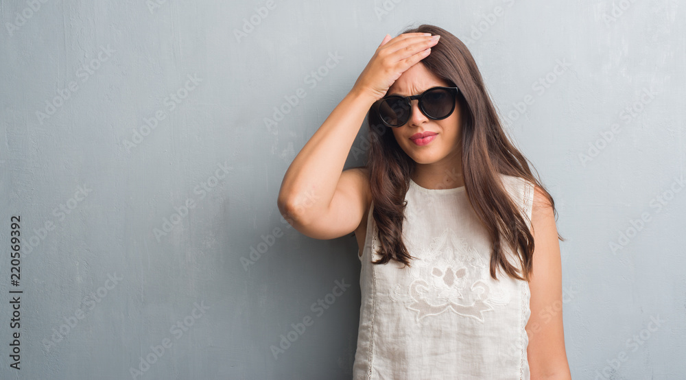 Young brunette woman over grunge grey wall wearing fashion sunglasses stressed with hand on head, shocked with shame and surprise face, angry and frustrated. Fear and upset for mistake.
