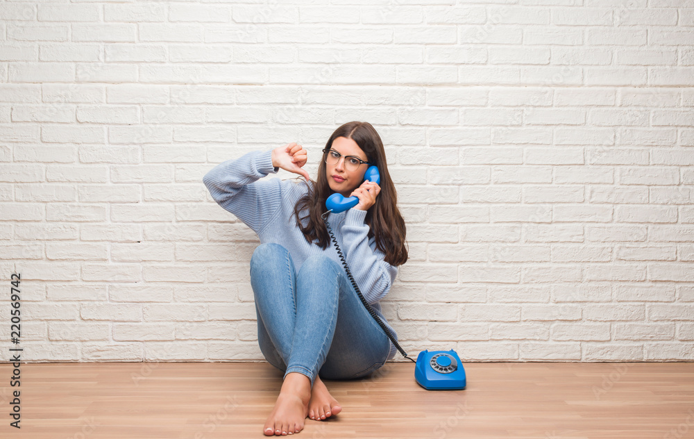 Young brunette woman sitting on the floor calling on vintage telephone with angry face, negative sign showing dislike with thumbs down, rejection concept Young brunette woman sitting on the floor calling on vintage telephone with angry face, negative sign showing dislike with thumbs down, rejection concept