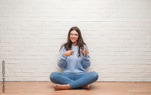 Young brunette woman sitting on the floor drinking glass of water very happy pointing with hand and finger