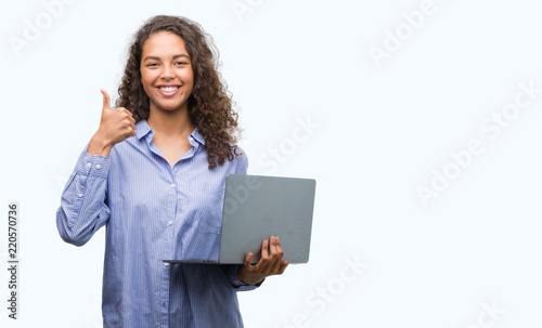 Young hispanic woman holding computer laptop happy with big smile doing ok sign, thumb up with fingers, excellent sign