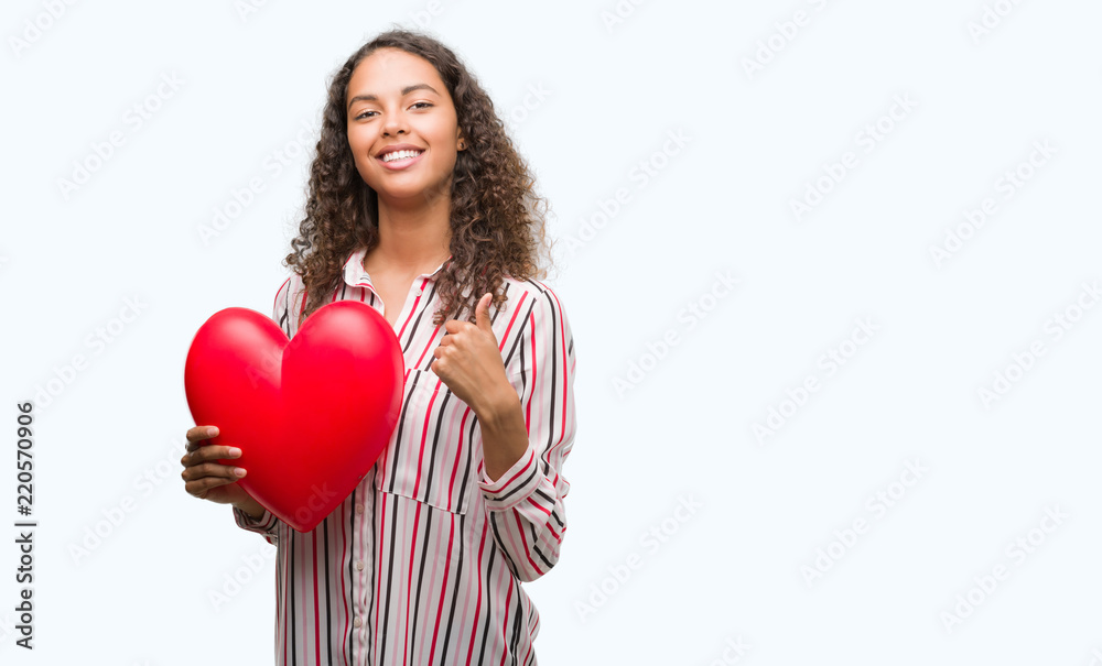 Young hispanic woman in love holding red heart happy with big smile doing ok sign, thumb up with fingers, excellent sign Young hispanic woman in love holding red heart happy with big smile doing ok sign, thumb up with fingers, excellent sign