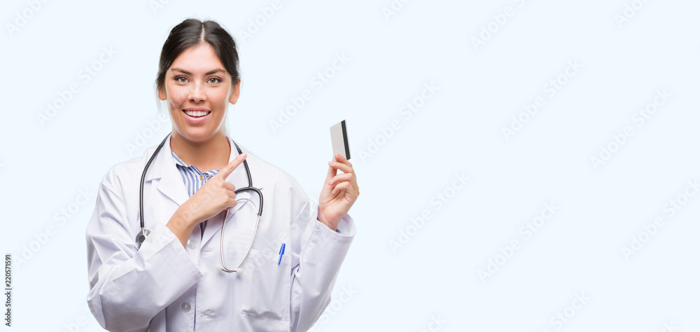 Young hispanic doctor woman holding credit card very happy pointing with hand and finger to the side Young hispanic doctor woman holding credit card very happy pointing with hand and finger to the side