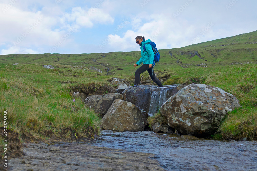 LOW ANGLE: Young female hikes over mountain stream flowing down rocky landscape.