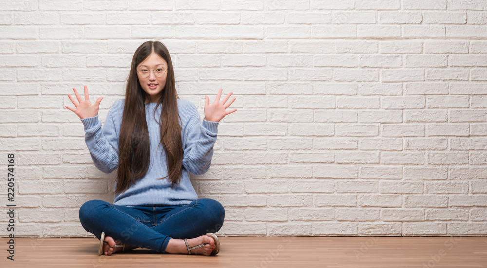 Young Chinese woman sitting on the floor over brick wall showing and pointing up with fingers number ten while smiling confident and happy. Young Chinese woman sitting on the floor over brick wall showing and pointing up with fingers number ten while smiling confident and happy.