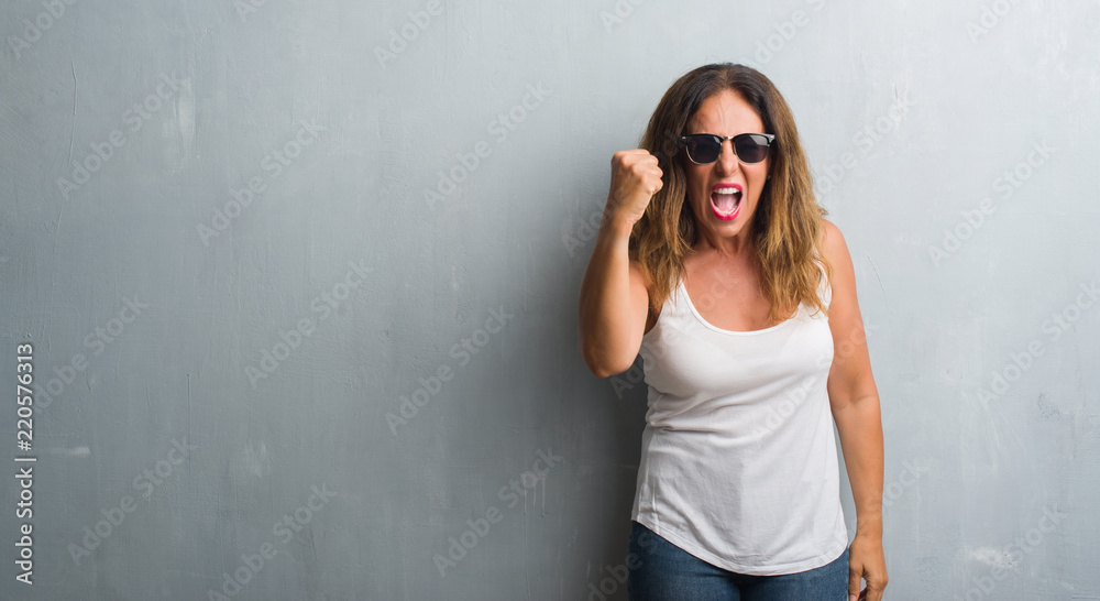 Middle age hispanic woman over grey wall wearing sunglasses angry and mad raising fist frustrated and furious while shouting with anger. Rage and aggressive concept. Middle age hispanic woman over grey wall wearing sunglasses angry and mad raising fist frustrated and furious while shouting with anger. Rage and aggressive concept.