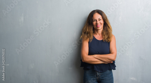 Middle age hispanic woman standing over grey grunge wall happy face smiling with crossed arms looking at the camera. Positive person.