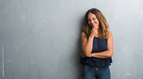 Middle age hispanic woman standing over grey grunge wall looking confident at the camera with smile with crossed arms and hand raised on chin. Thinking positive.