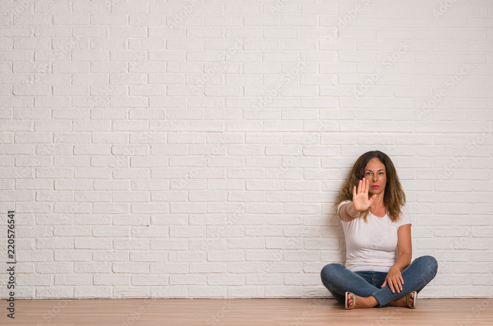 Middle age hispanic woman over white brick wall with open hand doing stop sign with serious and confident expression, defense gesture Middle age hispanic woman over white brick wall with open hand doing stop sign with serious and confident expression, defense gesture