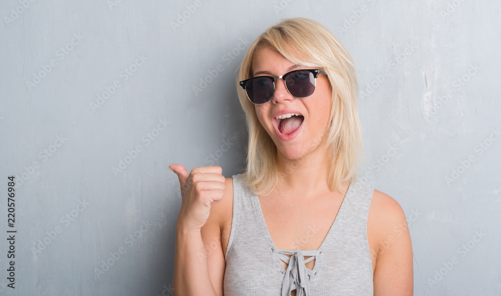 Adult caucasian woman over grunge grey wall wearing sunglasses pointing and showing with thumb up to the side with happy face smiling Adult caucasian woman over grunge grey wall wearing sunglasses pointing and showing with thumb up to the side with happy face smiling