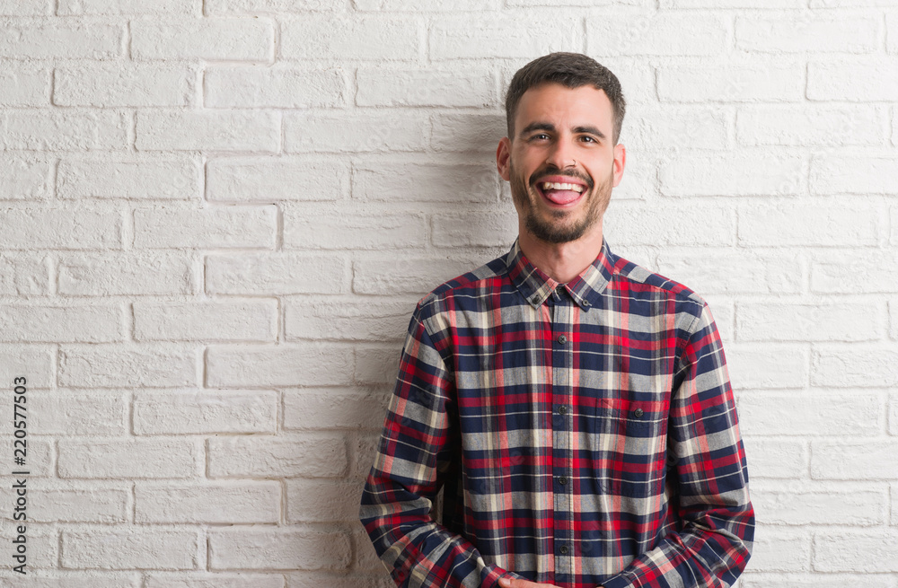 Young adult man standing over white brick wall sticking tongue out happy with funny expression. Emotion concept. Young adult man standing over white brick wall sticking tongue out happy with funny expression. Emotion concept.