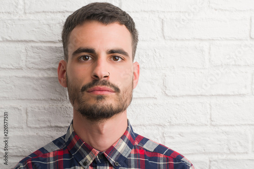 Young adult man standing over white brick wall with a confident expression on smart face thinking serious