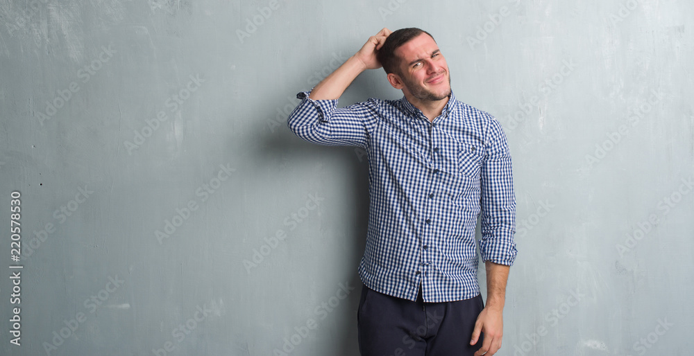 Young caucasian man over grey grunge wall confuse and wonder about question. Uncertain with doubt, thinking with hand on head. Pensive concept.