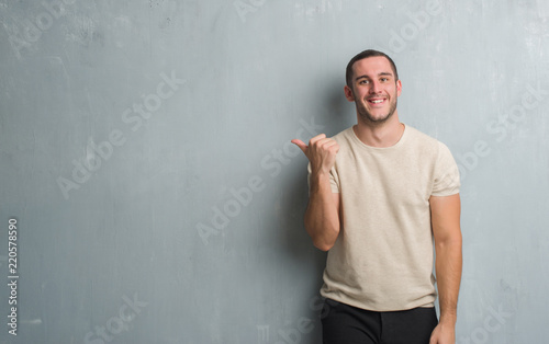 Young caucasian man over grey grunge wall smiling with happy face looking and pointing to the side with thumb up.