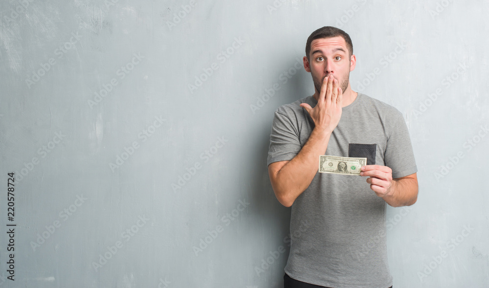 Young caucasian man over grey grunge wall showing one dollar cover mouth with hand shocked with shame for mistake, expression of fear, scared in silence, secret concept Young caucasian man over grey grunge wall showing one dollar cover mouth with hand shocked with shame for mistake, expression of fear, scared in silence, secret concept