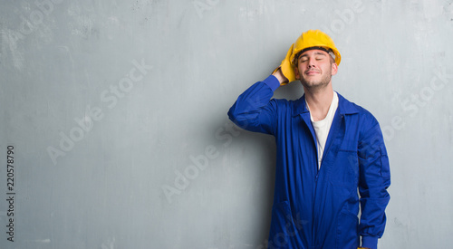 Young caucasian man over grey grunge wall wearing contractor uniform and safety helmet stressed with hand on head, shocked with shame and surprise face, angry and frustrated. Fear and upset