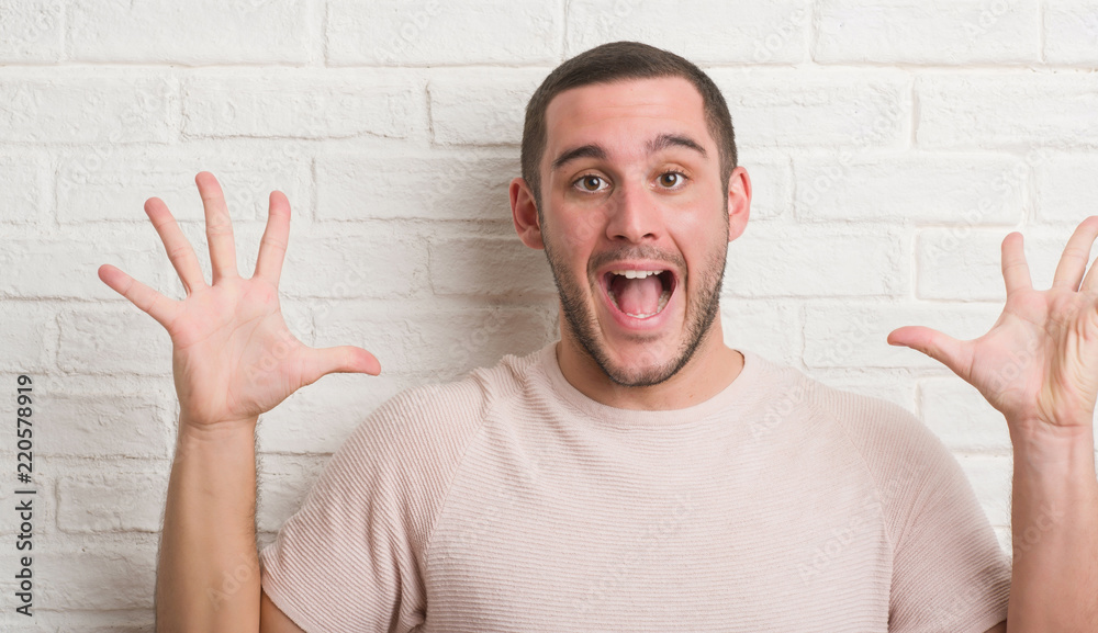 Young caucasian man standing over white brick wall very happy and excited, winner expression celebrating victory screaming with big smile and raised hands Young caucasian man standing over white brick wall very happy and excited, winner expression celebrating victory screaming with big smile and raised hands