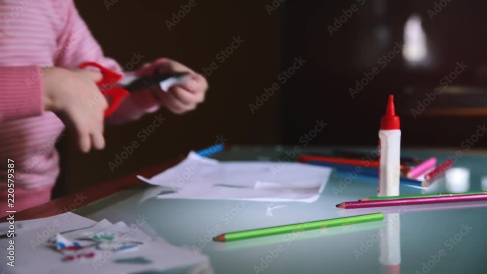 Camera sliding left on hands of little girl child in pink sweater cutting paper shapes at table with various stationery.