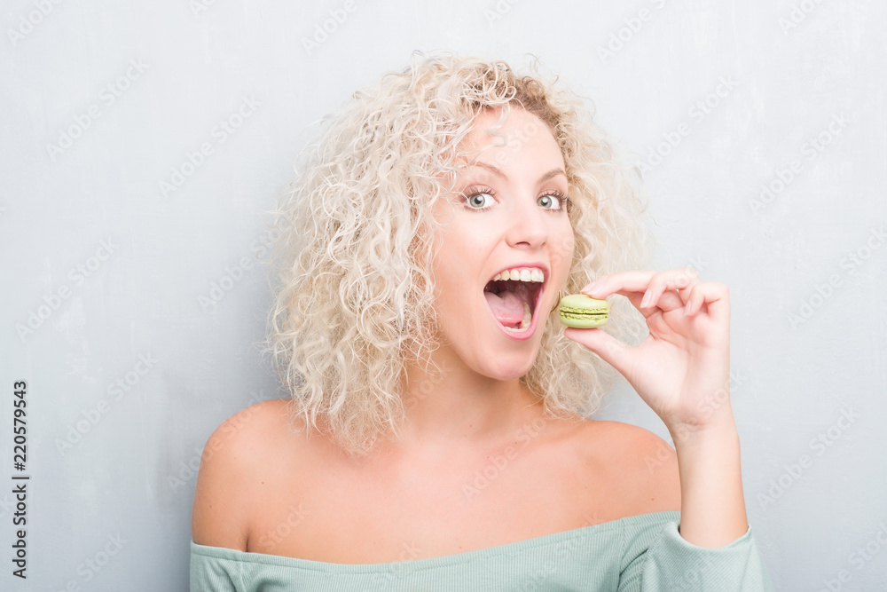Young blonde woman over grunge grey background eating macaron with a confident expression on smart face thinking serious Young blonde woman over grunge grey background eating macaron with a confident expression on smart face thinking serious