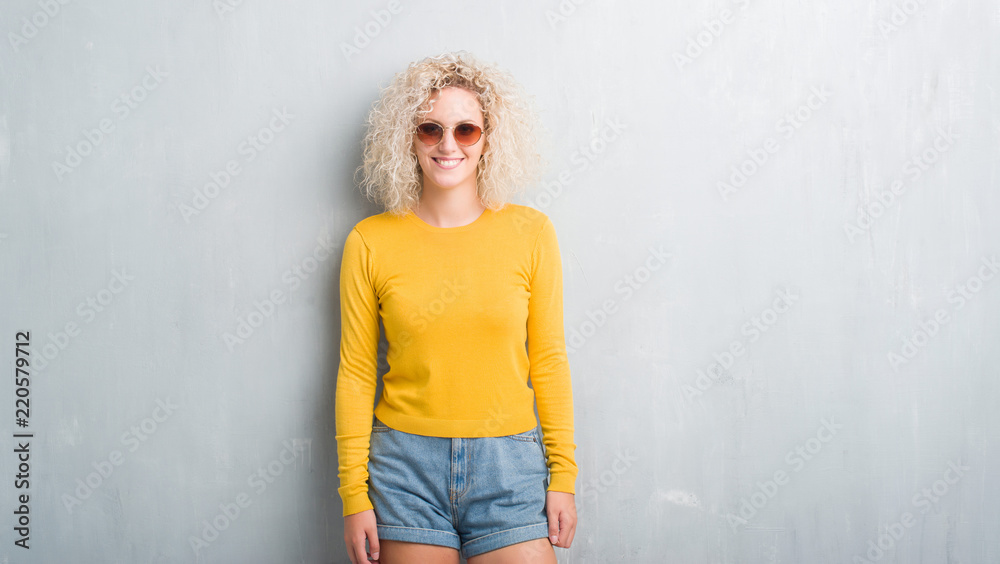 Young blonde woman with curly hair over grunge grey background with a happy and cool smile on face. Lucky person. Young blonde woman with curly hair over grunge grey background with a happy and cool smile on face. Lucky person.