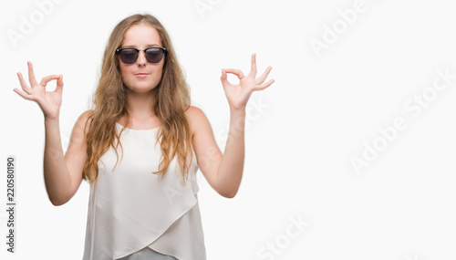 Young blonde woman wearing sunglasses relax and smiling with eyes closed doing meditation gesture with fingers. Yoga concept.
