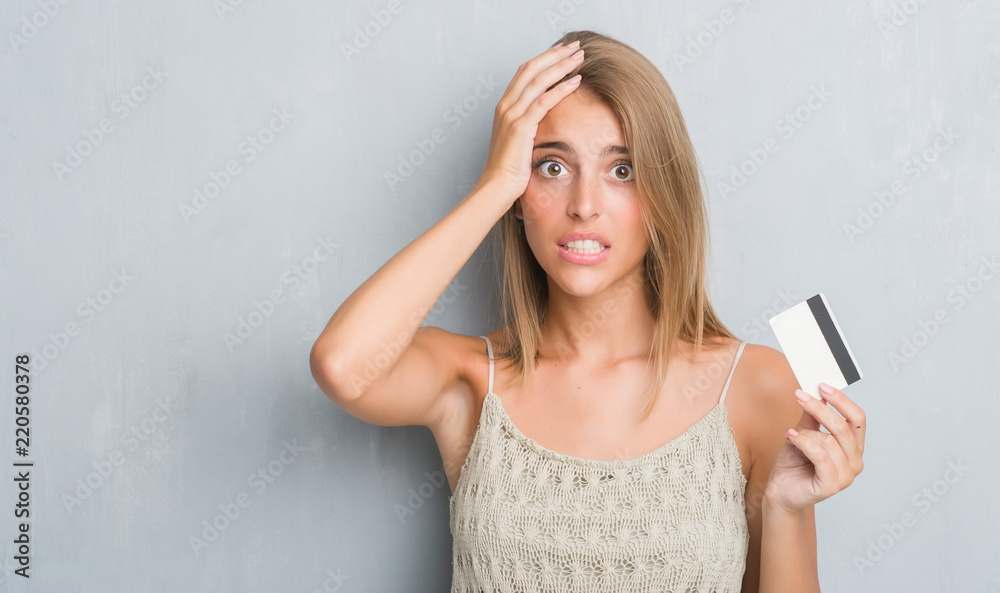 Beautiful young woman over grunge grey wall holding credit card stressed with hand on head, shocked with shame and surprise face, angry and frustrated. Fear and upset for mistake. Beautiful young woman over grunge grey wall holding credit card stressed with hand on head, shocked with shame and surprise face, angry and frustrated. Fear and upset for mistake.