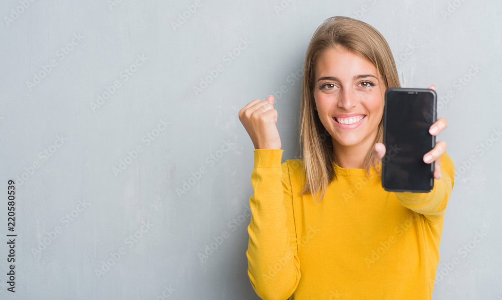 Fototapeta premium Beautiful young woman over grunge grey wall showing smartphone screen screaming proud and celebrating victory and success very excited, cheering emotion