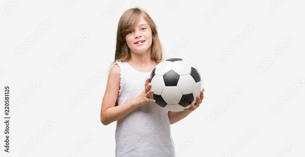 Young blonde toddler holding football ball with a happy face standing and smiling with a confident smile showing teeth
