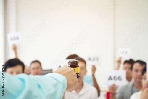 Close up auctioneer hand, holding gavel, wooden hammer, and blur group of people in auction room, one man raising hand up for bidding. Product or project auction market concept background photo