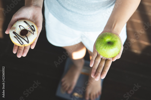 Choose a right choice for good health. Women is dieting comparison choice between donut and red apple during measuring weight on digital weight measure machine. photo