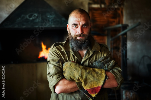 Portrait of a brutal, confident blacksmith, a man with a beard. Standing in the workshop, folding his arms over his chest.