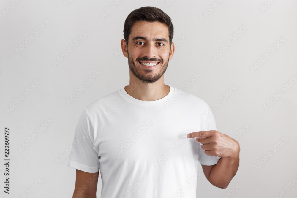 Horizontal shot of young handsome caucasian man isolated on gray background, smiling while pointing with index finger to white blank t-shirt, copyspace for advertising