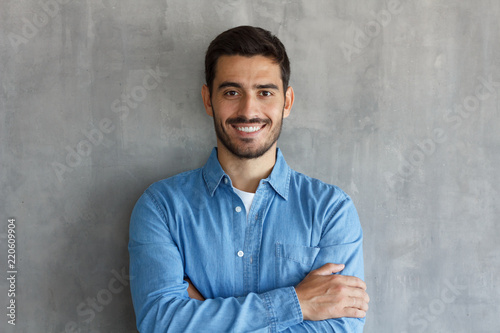 Smiling man in blue shirt, standing with crossed arms against gray textured wall photo