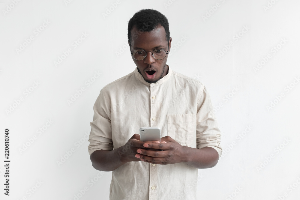 Portrait of handsome young african american man with mouth open, in bright shirt, holding smartphone, looking surprised and confused Portrait of handsome young african american man with mouth open, in bright shirt, holding smartphone, looking surprised and confused