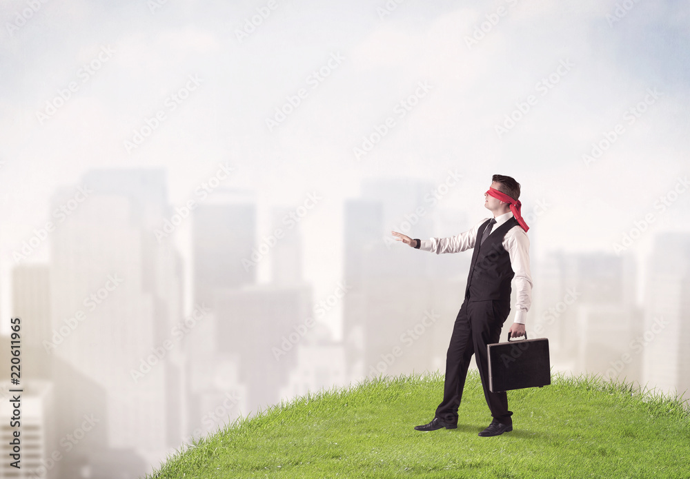 Young blindfolded businessman steps on a a patch of grass with a city in the background Young blindfolded businessman steps on a a patch of grass with a city in the background